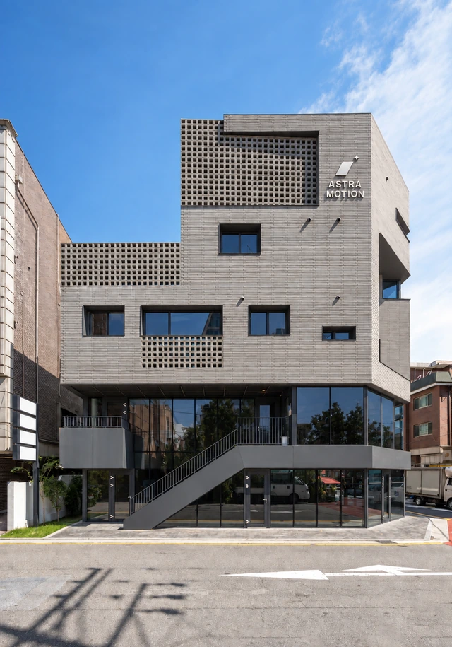 The ASTRA MOTION building: a grey brick building with perforated brick screens, a glass-fronted lower floor with an exterior staircase, and the ASTRA MOTION name and logo mark on its upper facade, under a blue sky.